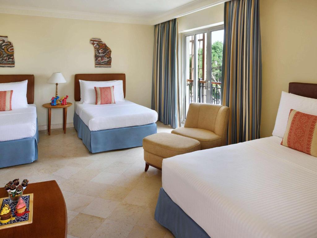 Liela viesnīcas numura iekārtojums ar divām atsevišķām gultām un atpūtas krēslu, gaiši krāsotiem aizkariem un dekoratīviem elementiem. Uz galda redzami saldumi un deserts.