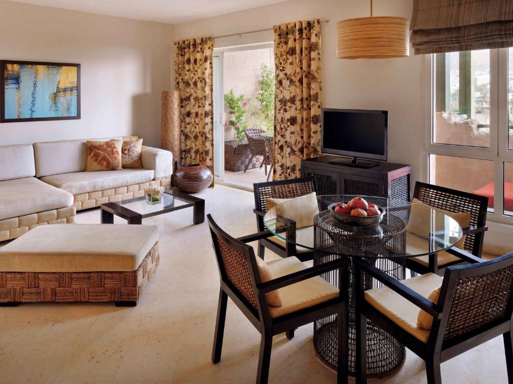 Mūsdienīgs viesistabas interjers ar dīvānu, stikla galdu un ērtām krēslām, ko papildina dekoratīvi akcenti un dabiska gaisma. Logi ved uz terasi ar zaļumiem.