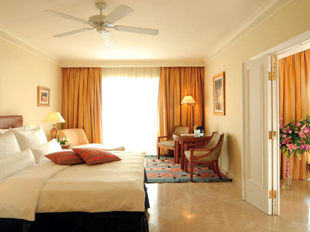 Lieliska viesnīcas istaba ar ērtu gultu, dīvānu un galdu, kas nodrošina komfortablu atmosfēru. Gaismas plūsmu papildina moderns ventilators un siltas krāsas aizkari.