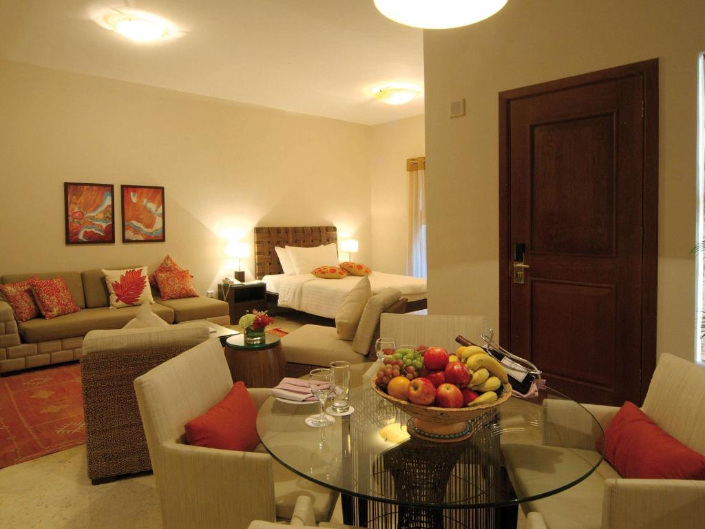 Mūsdienīgs viesnīcas numurs ar ērtu sēdvietu zonu un gultu, kur centrā atrodas stikla galds ar augļu trauku. Sienas rotā mākslas darbi, un telpa ir gaiša un viesmīlīga.