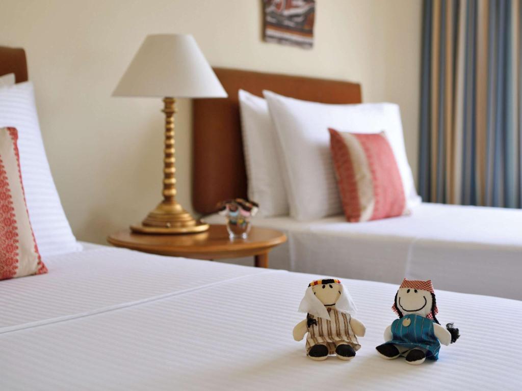 Divas rotaļlietu lelles guļamistabā uz gultas, blakus lampai un dekoratīvām spilveniem. Mūsdienīgs viesnīcas interjers ar vieglām krāsām un ērtu atmosfēru.