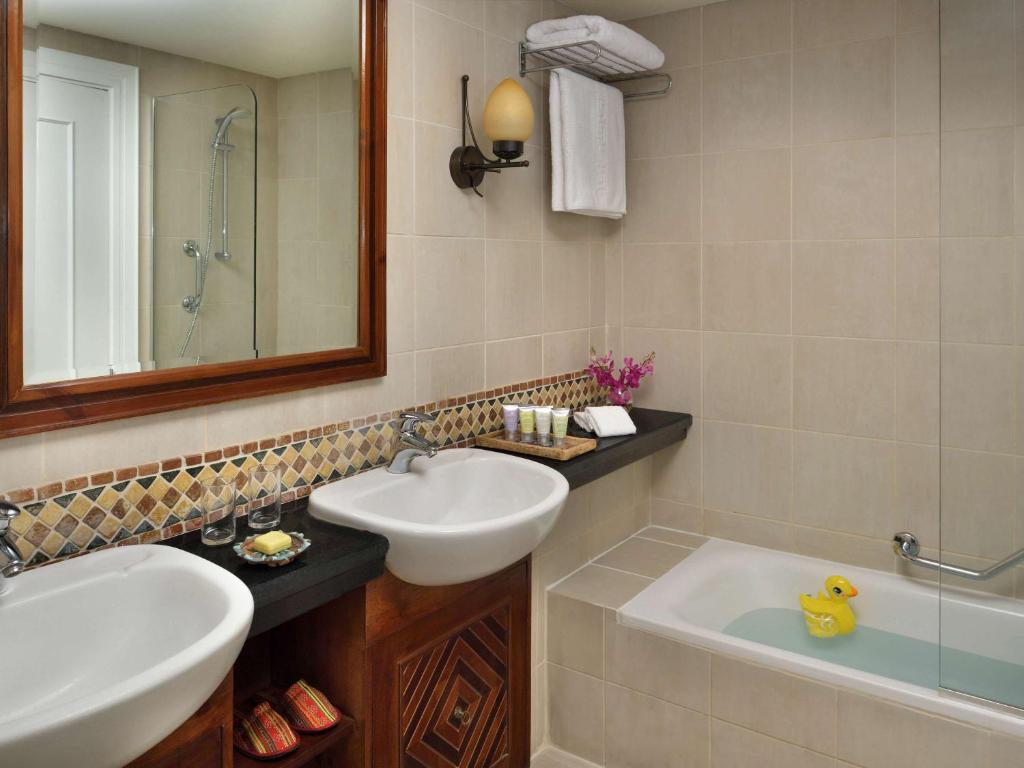 Mūsdienīgs vannas istabas interjers ar dubultu izlietni, dušas kabīni un vannu, kurā peld dzeltenais pīlēns. Uz letes ir izvietoti higiēnas piederumi un ziedi.