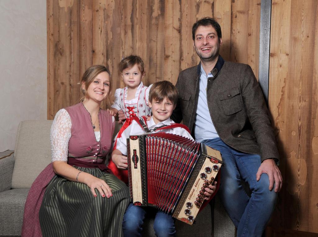 Ģimenes portrets ar bērniem, kurā ir četri cilvēki, no kuriem divi tur akordeonu. Fons ir koka siena, un visi ir tērpušies tradicionālajos apģērbos.