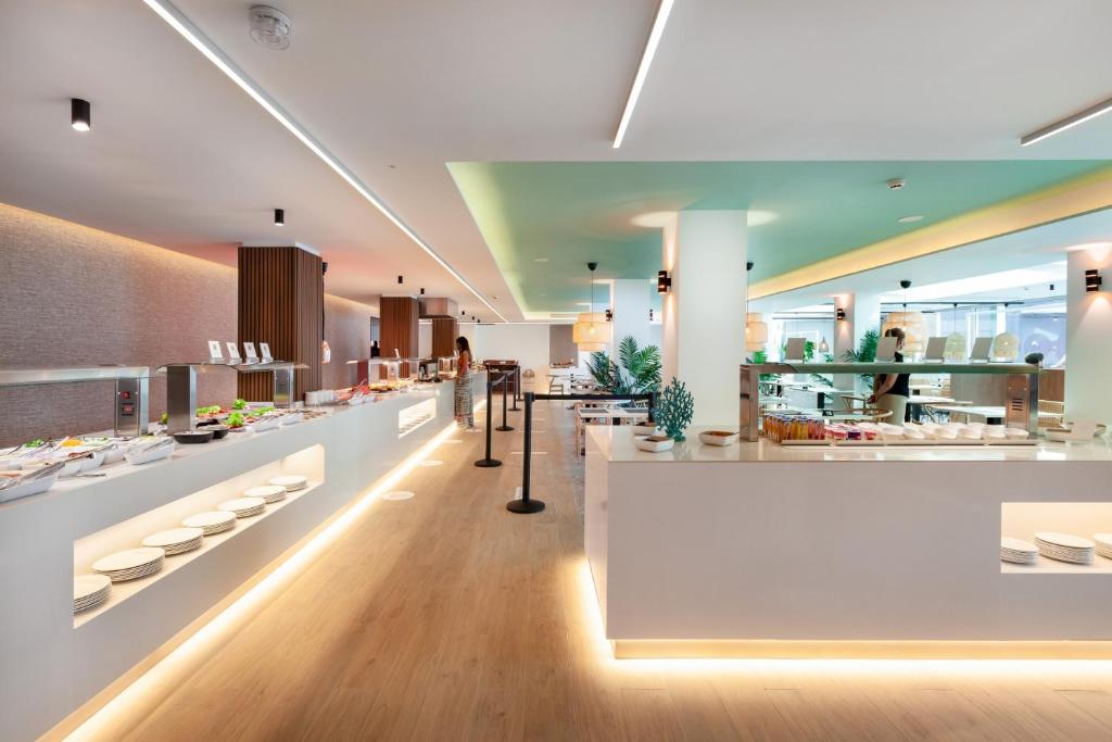 Mūsdienīgs restorāns ar plašu bufeti, kurā redzamas dažādas ēdienu izvēles un elegantas interjera detaļas. Apgaismojums un augu dekorācijas piešķir siltu un viesmīlīgu atmosfēru.