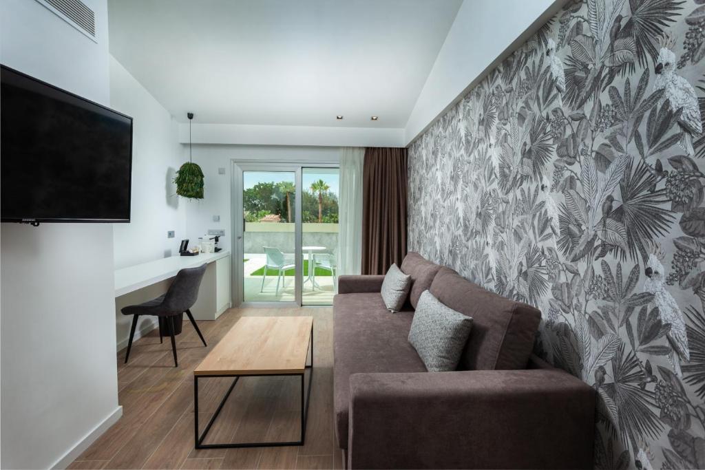 Mūsdienīgs viesnīcas numurs ar dīvānu, koka galdu un televizoru, kas piedāvā piekļuvi terasei ar skatu uz dārzu. Interjers ir papildināts ar augu motīviem uz sienas.