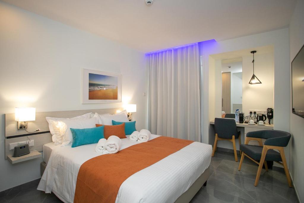 Mūsdienīga viesnīcas numura iekārtojums ar ērtu gultu, spilveniem un akcentiem oranžā un zilo krāsā, kā arī darba vieta ar krēslu un kafijas pagatavošanas iespējām.