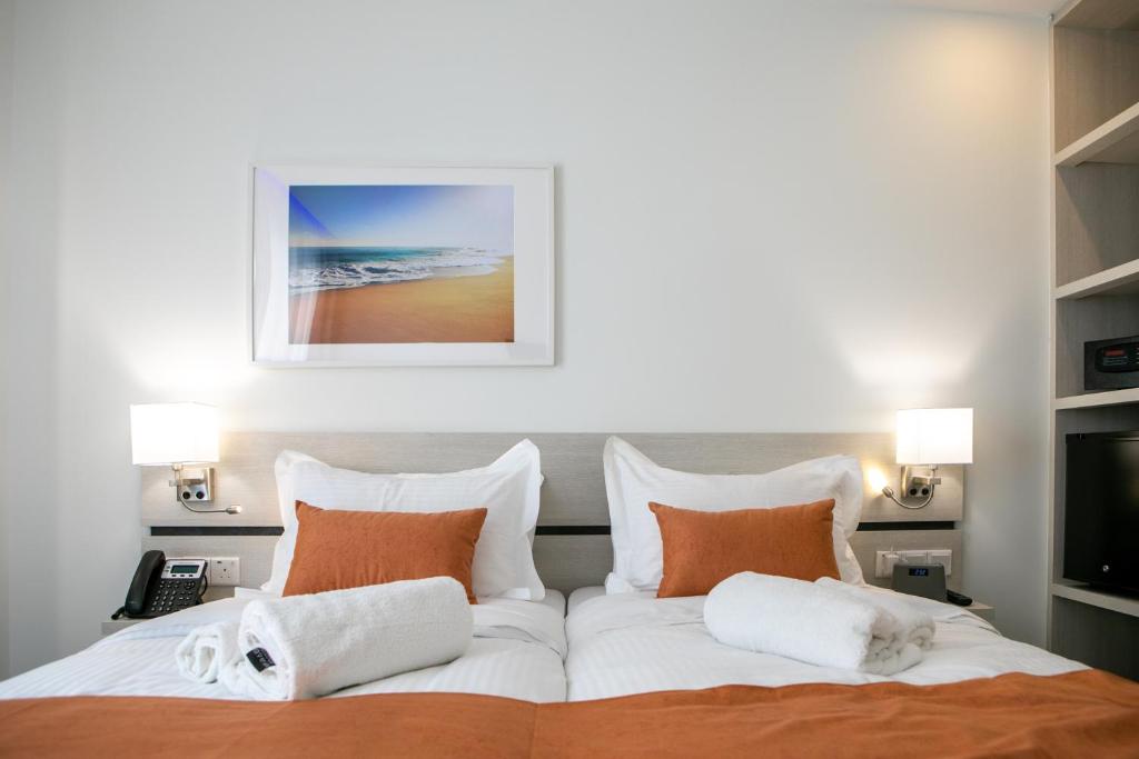 Mūsdienīgs viesnīcas numurs ar divām gultām, oranžiem spilveniem un baltām dvieļiem, kā arī pie sienas piekarinātu gleznas attēlu ar pludmali. Apgaismojums un mūsdienīgas mēbeles rada ērtu atmosfēru.