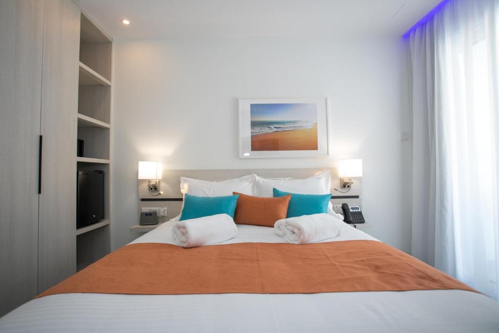 Moderns viesnīcas numurs ar ērtu gultu, spilveniem un gaismu, kas rada relaksējošu atmosfēru. Interjerā redzama plauktu sistēma un mākslas darbs ar pludmales ainavu.