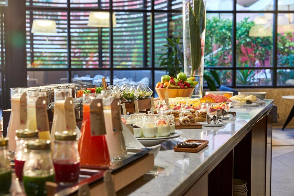 Bagātīgs brokastu bufete ar dažādiem dzērieniem un ēdieniem, tostarp augļiem, saldumiem un salātiem, ierīkošana gaišā un modernā vidē.