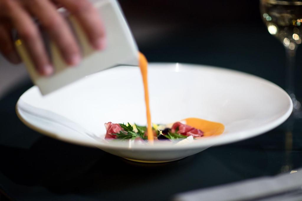 Gourmet ēdiens tiek pasniegts ar krāsainu mērci, kas tiek ielej taisni uz šķīvja, radot estētisku un izsmalcinātu izskatu. Fons ir tumšs un elegants, papildināts ar vīna glāzi.