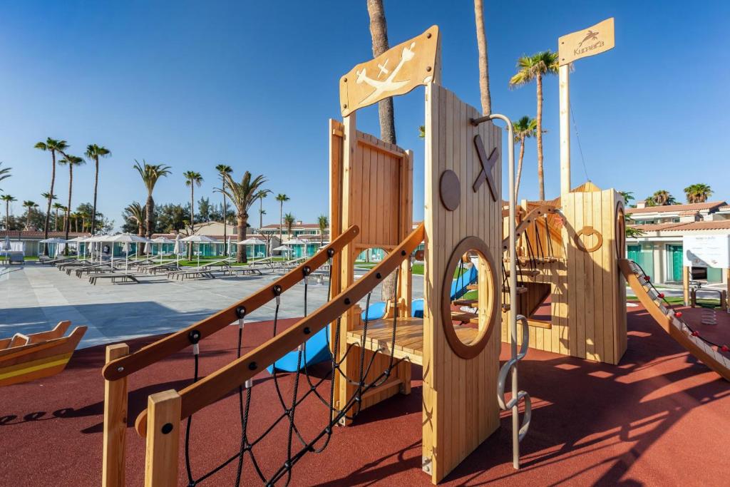 Koka bērnu spēļu laukums ar dažādām kāpšanas struktūrām un slidkalniņiem, apjozts ar palmām un saulainu debesi. Ideāla vieta bērnu izklaidei un aktivitātēm.