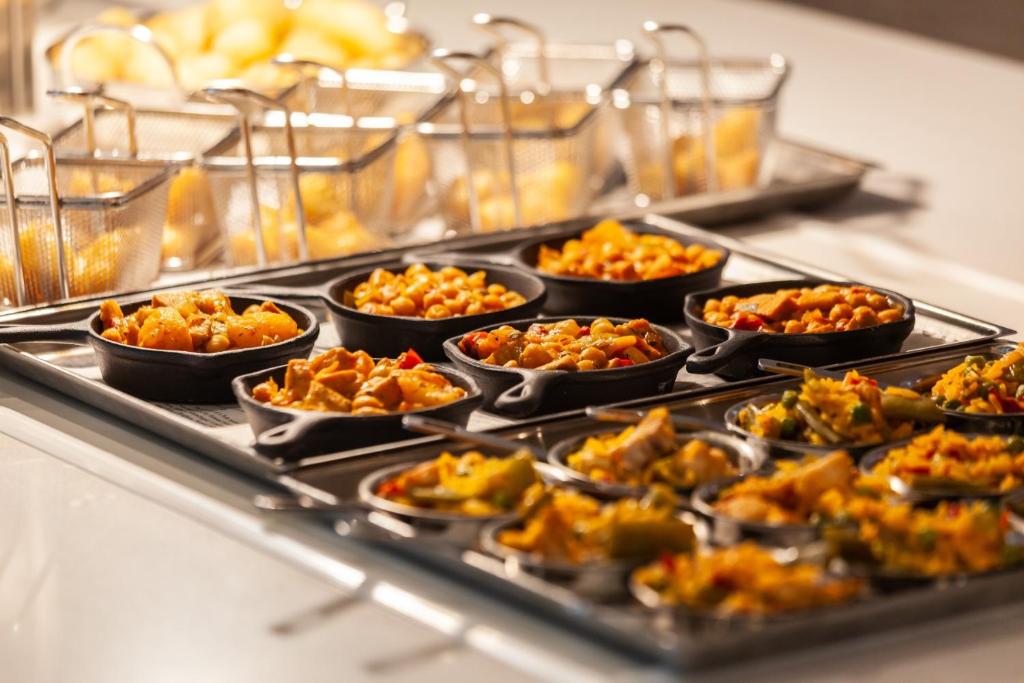 Dažādu ēdienu pasniegšana mini pannās, ietverot dārzeņu un pākšaugu ēdienus, ar fona groziem ceptiem gardumiem.