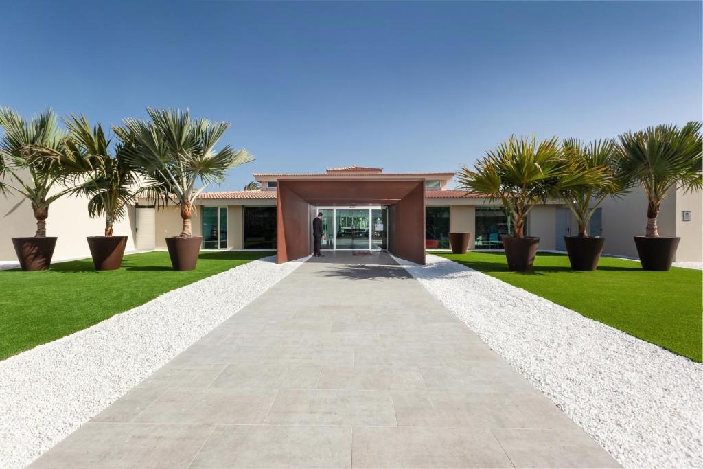 Moderns arhitektūras dizains ar elegantu ieeju, kurā redzami lieli podi ar palmām un zaļš zālājs, veido pievilcīgu ainavu. Ieeja ir veidota no metāla un stikla, radot mūsdienīgu atmosfēru.