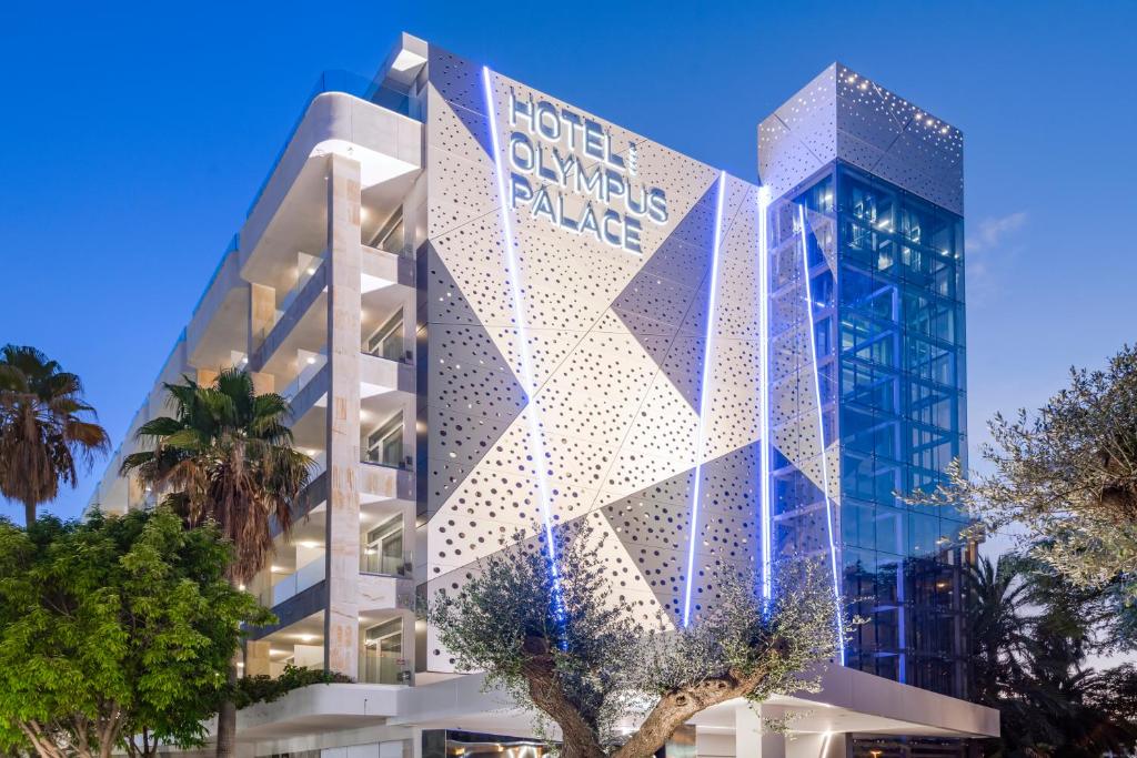 Moderns viesnīcas ēkas dizains ar izteiksmīgu apgaismojumu, kas izceļ "Hotel Olympus Palace" nosaukumu. Apkārtne ir zaļa ar palmām un citiem kokiem.