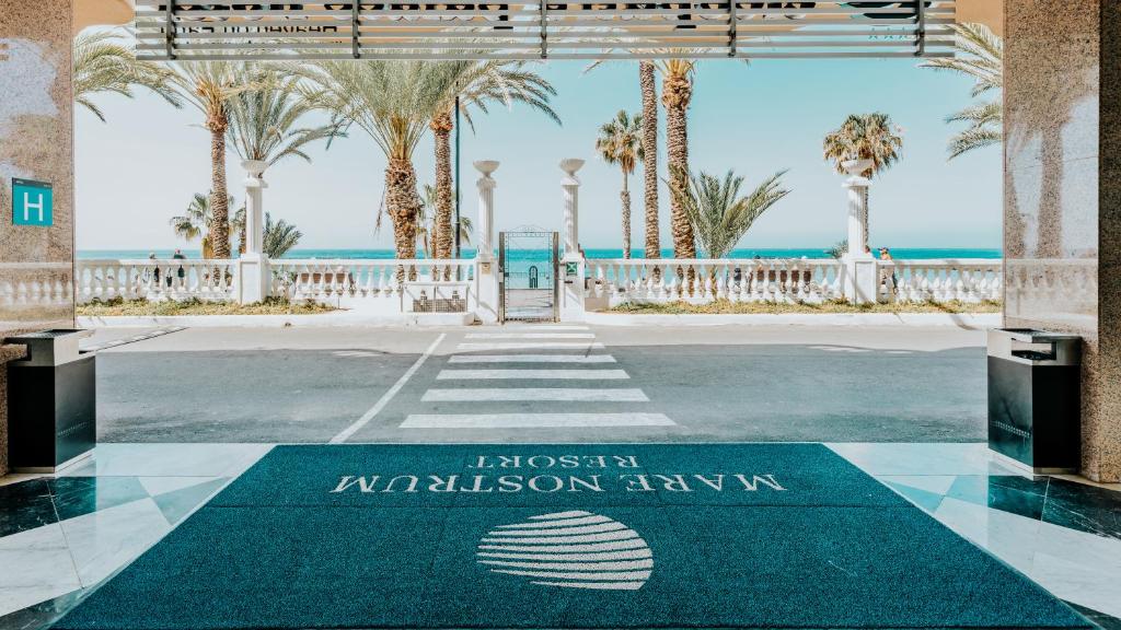 Viesnīcas ieeja ar skatu uz pludmali un palmām, kas rada relaksējošu atmosfēru. Redzams viesnīcas logotips uz grīdas paklājiņa.
