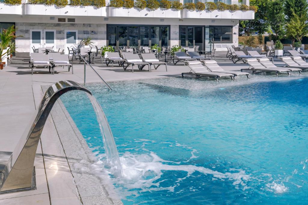 Mūsdienīgs peldbaseins ar plūstošu ūdens strūklu un sauļošanās krēsliem gar malām, ideāla vieta atpūtai. Skats uz ēku ar lieliem logiem un zaļām augiem.