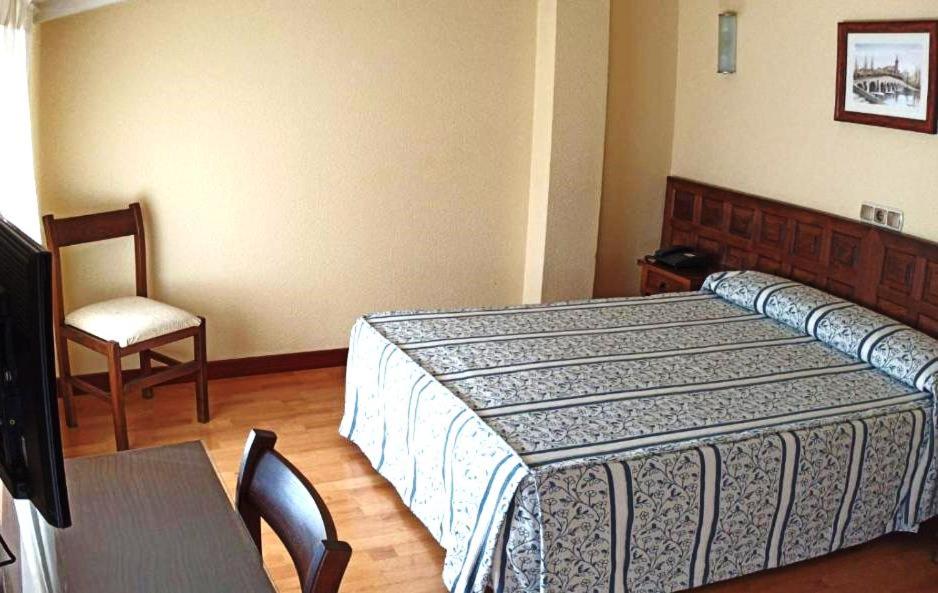 Vienkārša viesnīcas numura interjers ar gultu, koka krēslu un televizoru, kas atrodas uz koka grīdas. Numurā ir gaišas sienas un gaisma, kas ieplūst caur logu.