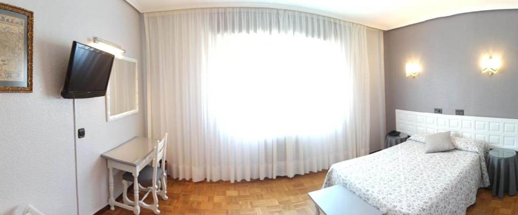 Gaiša viesnīcas istaba ar gultu, rakstāmgaldu un televizoru pie sienas, lieli logi ar aizkariem un koka grīda.