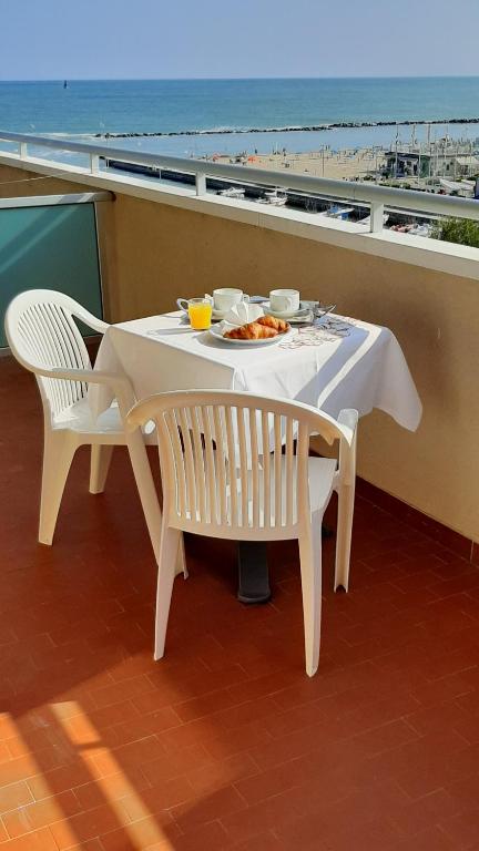Balkona skats uz jūru ar galdu, kurā ir brokastu komplekts: kafija, oranžsula un kruasāns. Fons ietver pludmali un jūru, radot relaksējošu atmosfēru.