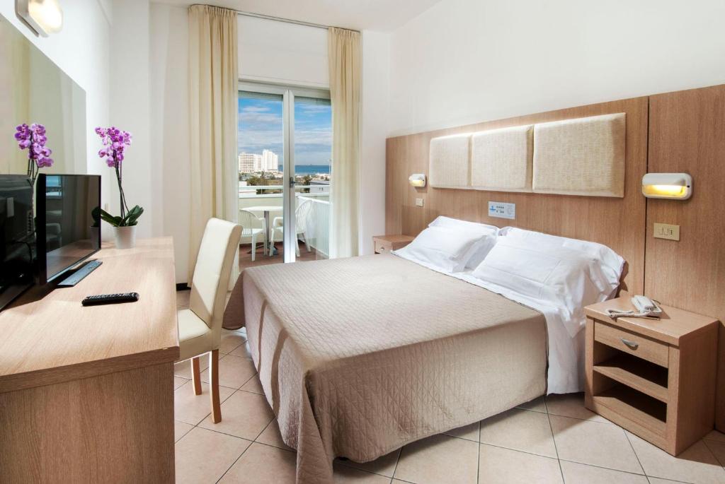 Mūsdienīga viesnīcas numura iekārtojums ar gultu, televizoru un rakstāmgaldu. Logā redzams skats uz terasi un jūru.