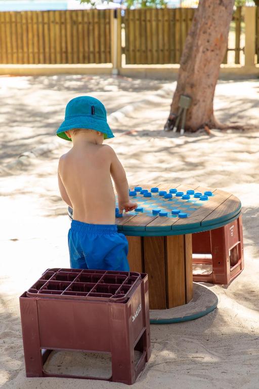 Bērns, tērpies zilos šortos un zaļā cepurē, spēlē pie galda ar krāsainām figūrām pludmales vidē. Fons ir smilšains, ar koka žogu un koku.