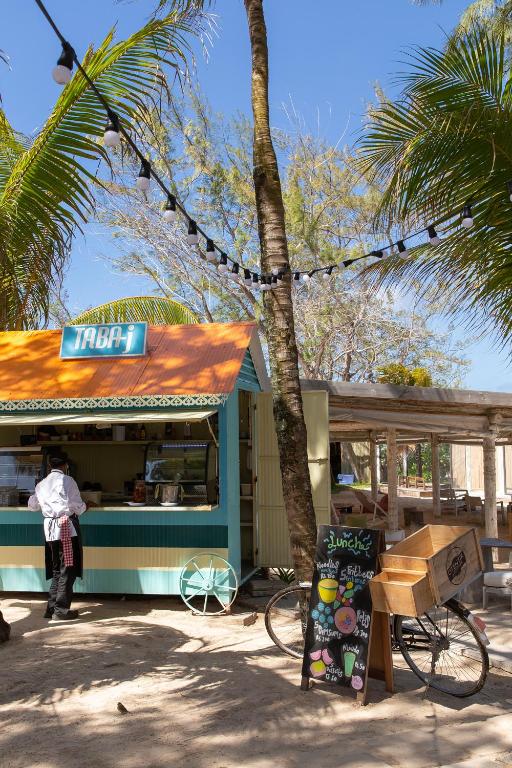 Kafejnīca ar nosaukumu "TABA-i", kas atrodas tropiskā vidē, ar palmām un dekoratīviem gaismas virknēm. Apmeklētājs pasūta ēdienu, bet blakus ir velosipēds un informācijas tāfele ar piedāvājumiem.