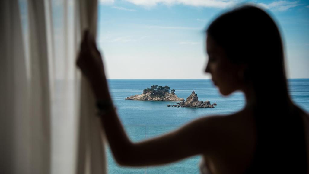Skats no loga uz klusu jūru un nelielu salu, bet priekšplānā ir sievietes siluets, kas velk aizkari. Ideāla ainava relaksācijai un mieram.