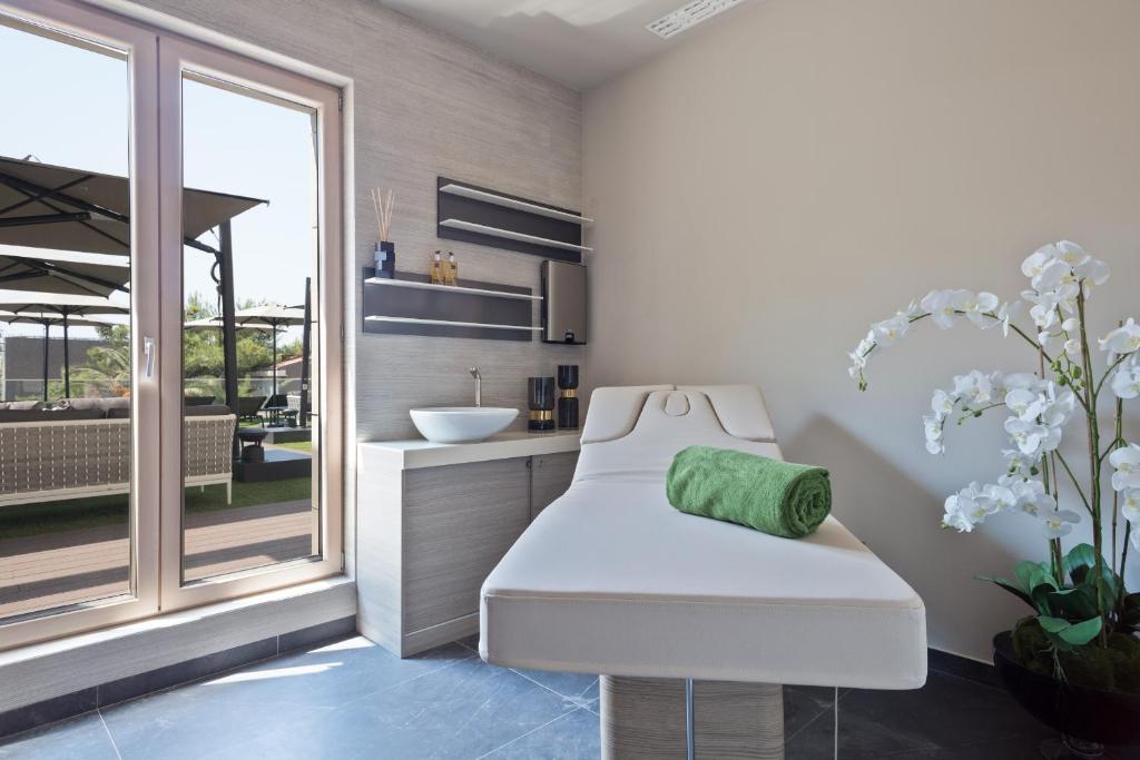 Mūsdienīgs spa telpa ar masāžas galdu, zaļu dvieli un dekoratīvām orhidejām, kas nodrošina relaksējošu atmosfēru. Lieli logi ļauj ieplūst dabiskai gaismai.