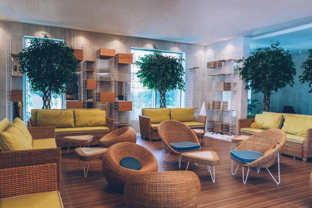 Mūsdienīga atpūtas telpa ar zaļām dīvānu mēbelēm un rotājošām koka elementiem, kas rada relaksējošu atmosfēru. Telpā ir lieli augi un dabīga apgaismojuma avoti.