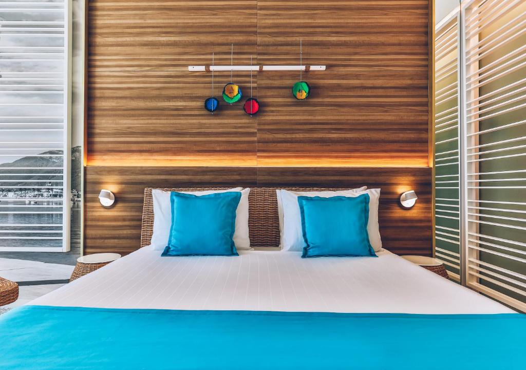 Mūsdienīga guļamistaba ar koka sienu un spilgtiem akcentiem, izceļot zilu gultas pārklāju un spilvenus. Interjeru papildina dekoratīvi elementi un gaismas avoti, radot viesmīlīgu atmosfēru.