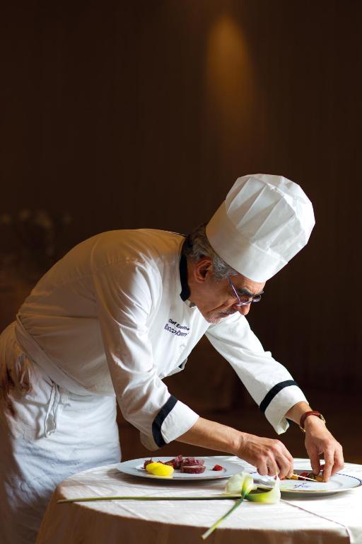 Pavārs, kas rūpīgi dekorē ēdienu uz galda, izmantojot svaigus sastāvdaļas. Fons ir neitrāls un izceltas gaismas.