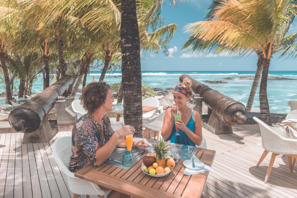 Tropu restorāns pie okeāna, kur divi cilvēki bauda kokteiļus un augļus, apkārt palmām un skaistām ainavām.