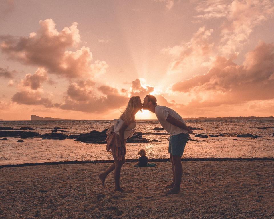 Romantisks skats uz pāri, kas skūpstās pludmalē saulrieta gaismā, ar jūru un mākoņiem fonā.