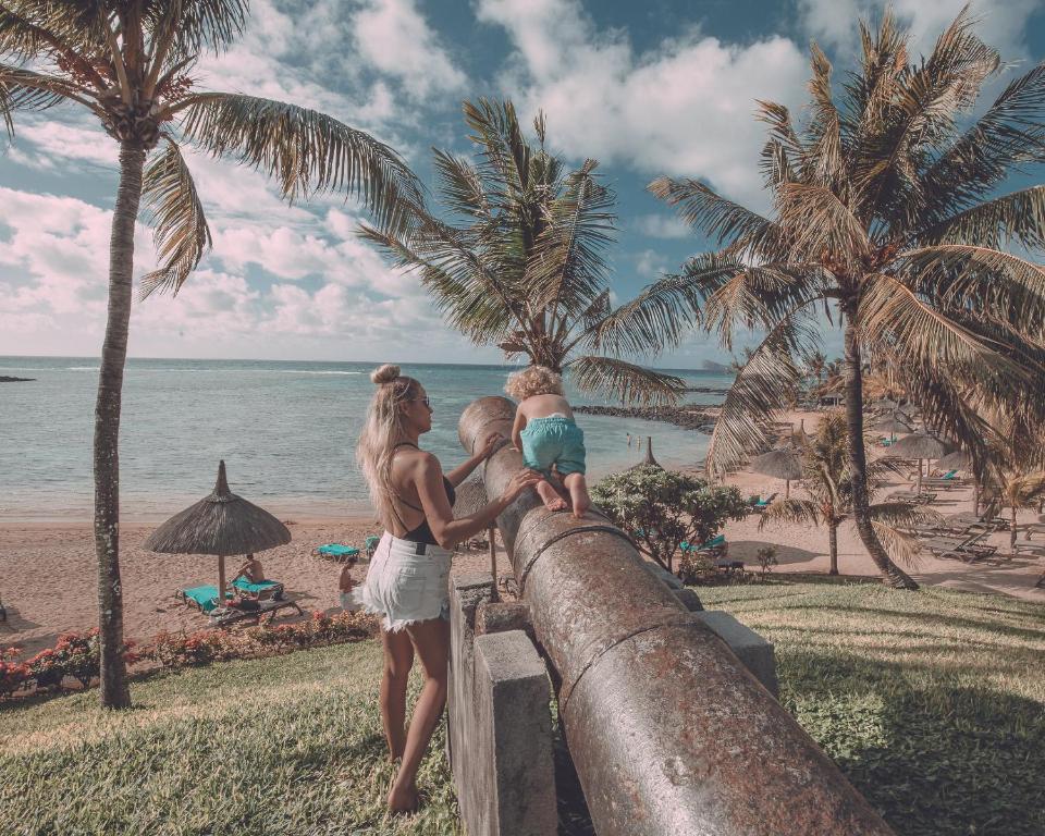 Mākslas darbs ar sievieti un bērnu, kas izpēta vēsturisku kanonu pludmalē, ko ieskauj palmu koki un skaista jūras ainava.