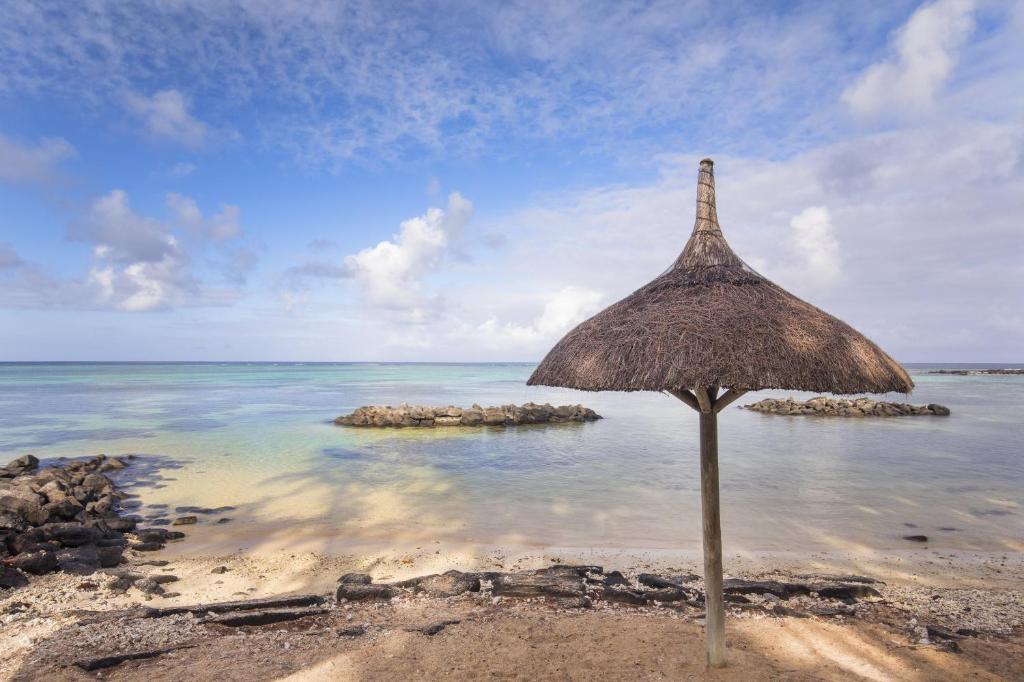 Pludmales ainava ar salmu nojumeņu, kas sniedzas virs smilšainās krasta, ar mierīgu ūdeni un akmeņu veidojumiem fona.