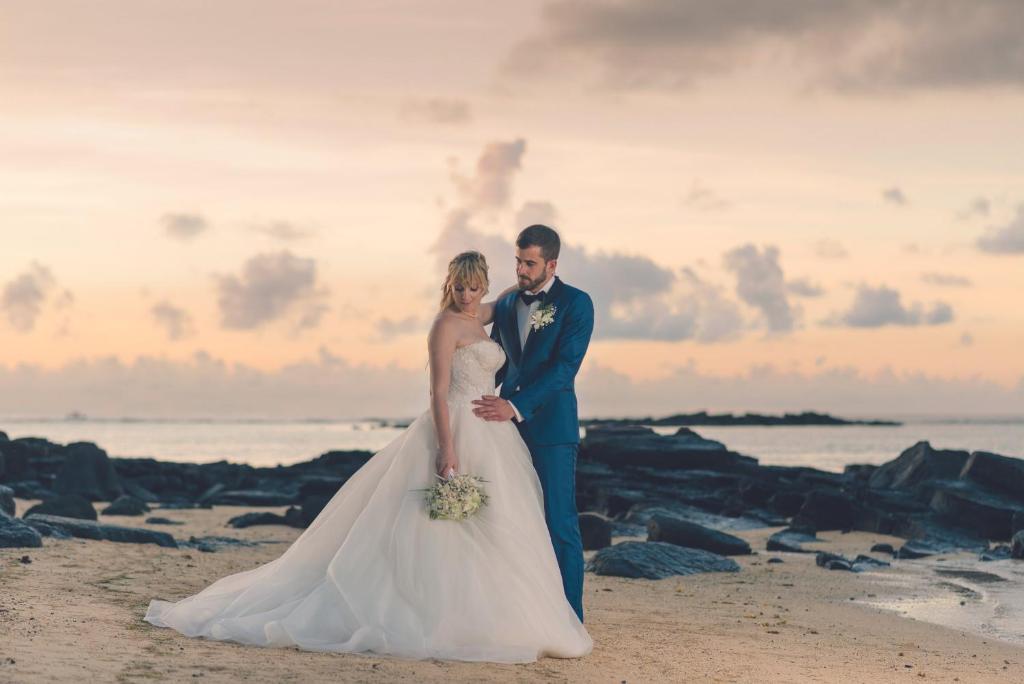 Jaunlaulātie stāv pludmalē, tērpti elegantiem kāzu tērpiem, ar saulrietu fona un akmeņainu krastu.