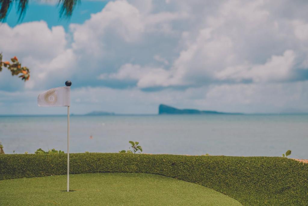 Zaļš golfa laukums ar mazu baltu karodziņu, kas izvirzīts jūras krastā, kam fonā redzama skaista ainava ar mākoņainu debesi un attālu salu.