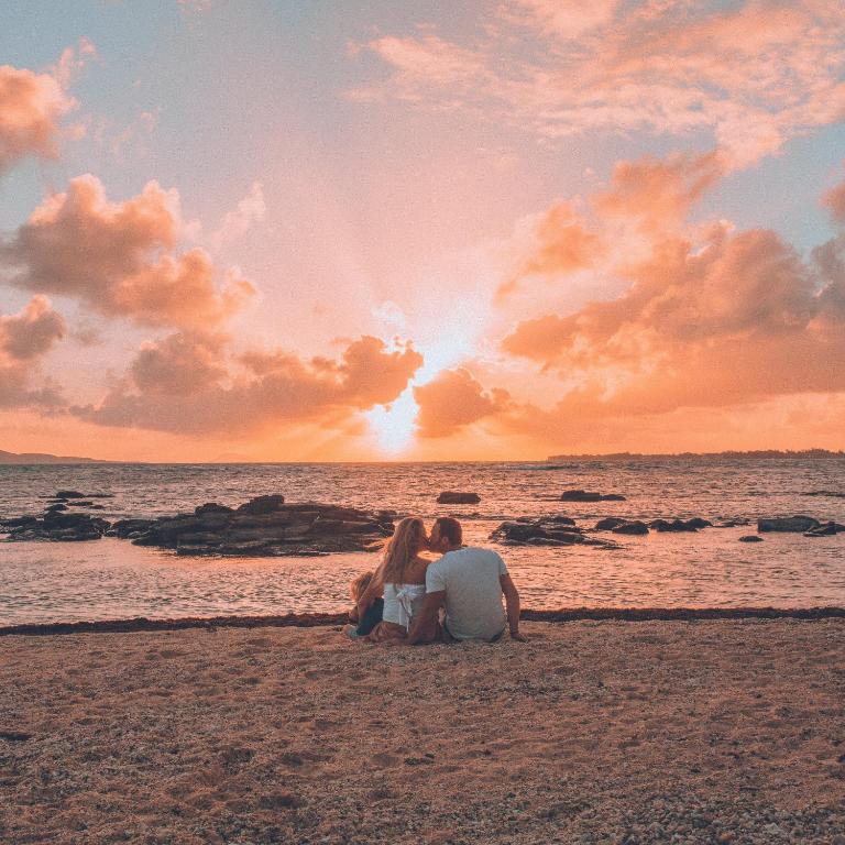 Pāris sēž uz pludmales, skatoties saulrietā, kad debesīs ir rozā un oranžs apgaismojums. Apkārt ir akmeņi un viļņi, radot romantisku noskaņu.