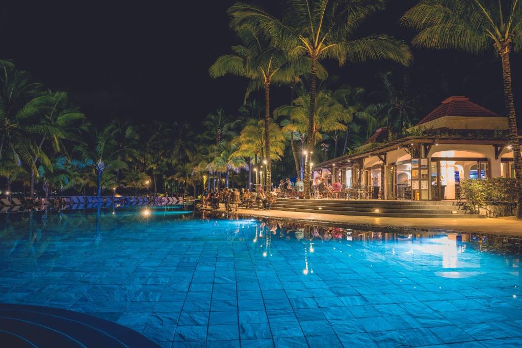 Nakts ainava ar spožu baseinu un tropiskiem palmu kokiem. Viesnīcas terase ir apgaismota, un apmeklētāji bauda atpūtu pie ūdens.