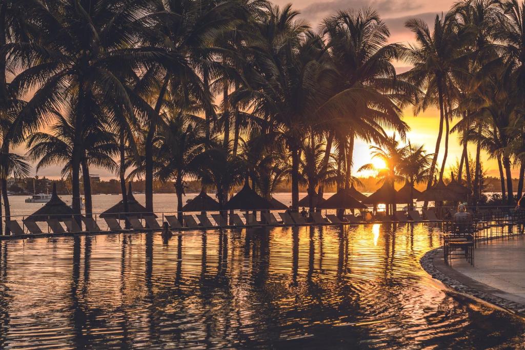 Saulriets aiz palmu silueta pie baseina, atspoguļojot mierīgu ūdens virsmu un siltas krāsas debesīs. Ideāla atpūtas atmosfēra tropiskā vidē.