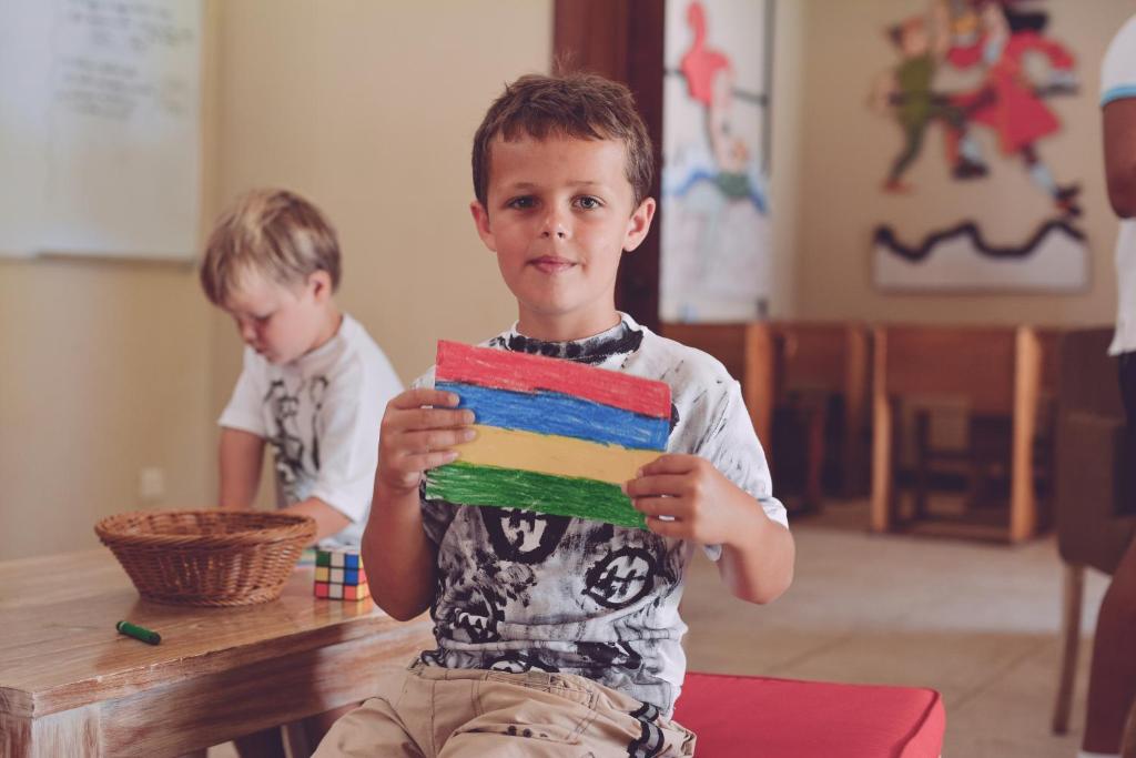Bērns, kas tur krāsotu karogu ar sarkanu, zilu un zaļu krāsu, sēž mākslinieciskā vidē, kamēr cits bērns spēlējas blakus.