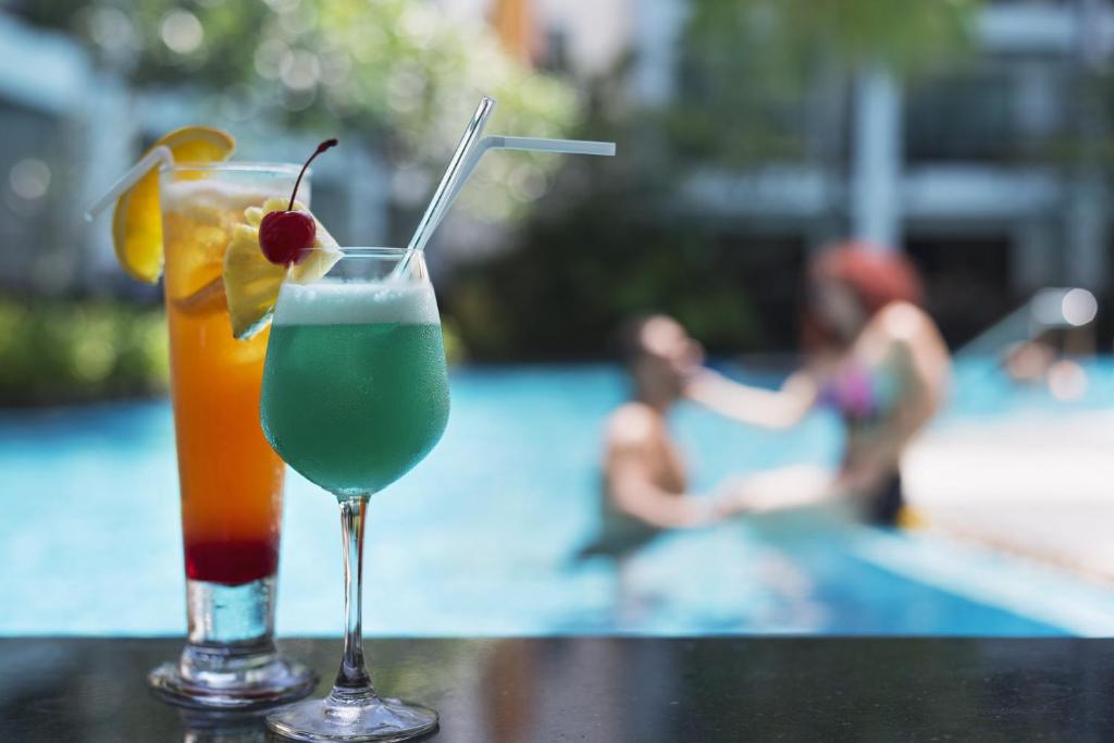 Divi krāsaini kokteiļi, viens ar apelsīnu un ķiršu dekoru, otrs zaļš un putojošs, atrodas uz galda, kamēr pāris izbauda brīvdienas baseinā.