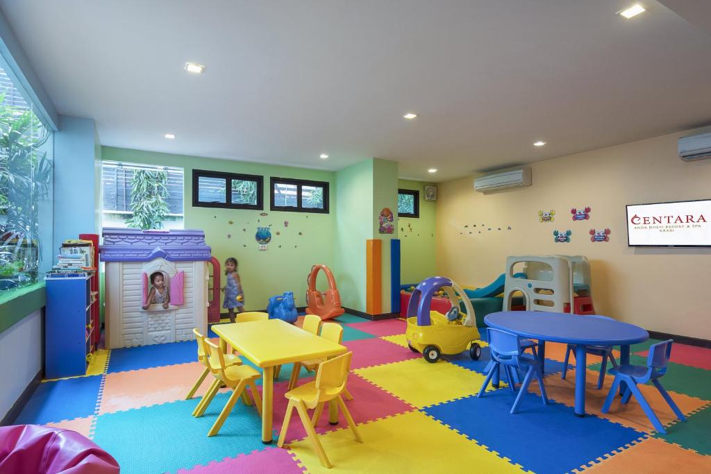 Krāsaina bērnu spēļu istaba ar plastmasas mēbelēm, rotaļu māju un dažādiem rotaļlietu elementiem. Telpā ir arī bērnu galdi un krēsli, kā arī televizors ar logo.