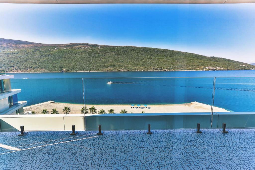 Skats uz mierīgu jūru un kalnu ainavu no modernā balkona, kur redzama pludmale ar palmām un saulessargiem. Ideāla vieta atpūtai pie ūdens.