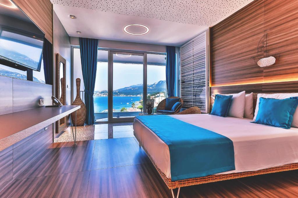 Mūsdienīgs viesnīcas numurs ar skatu uz kalniem un jūru, iekārtots ar gaišām krāsām un komfortablu mēbelējumu. Numurā ir liels gulta ar zilu gultas pārklāju un televizors pie sienas.