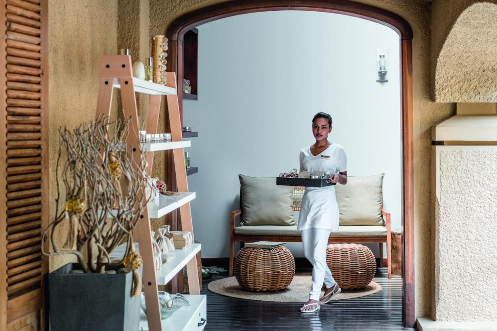 Skaista un eleganta viesmīle, tērpta baltā apģērbā, ienāk telpā ar pasniegšanas paplāti, fonā redzama moderna interjera dizaina iekārtojums.