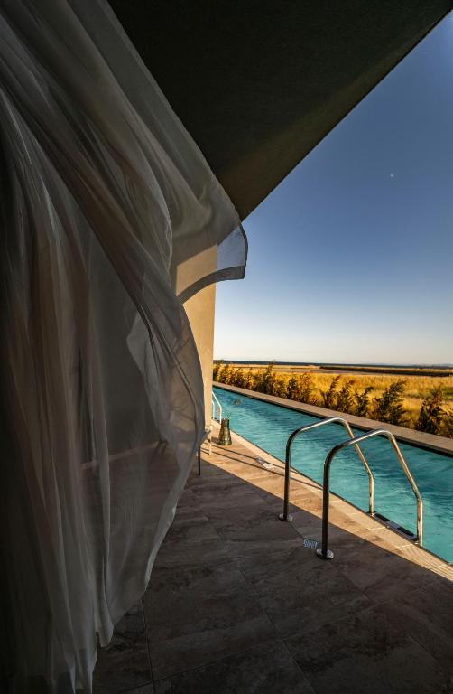 Skats uz baseinu no terases, kur gaisīga aizkaru drapērija plūst vēja ietekmē. Fons ar zilo debesu un zāles ainavu rada mierīgu atmosfēru.