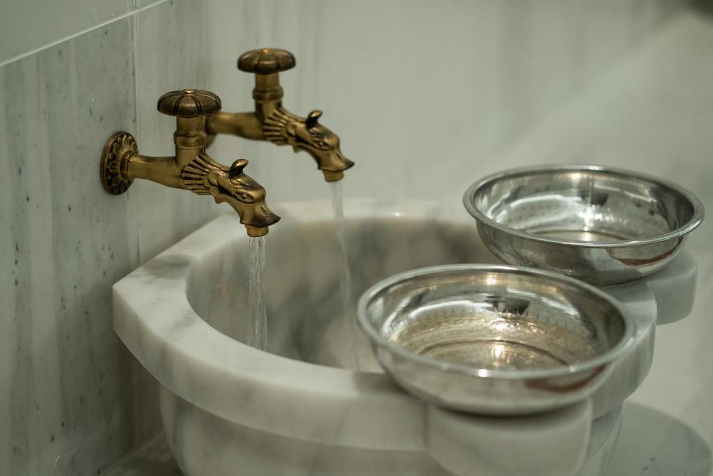 Divi vintage stila krāni ar ūdeni, kas plūst no zelta krāsu izlietnes, blakus divām metāla bļodām.