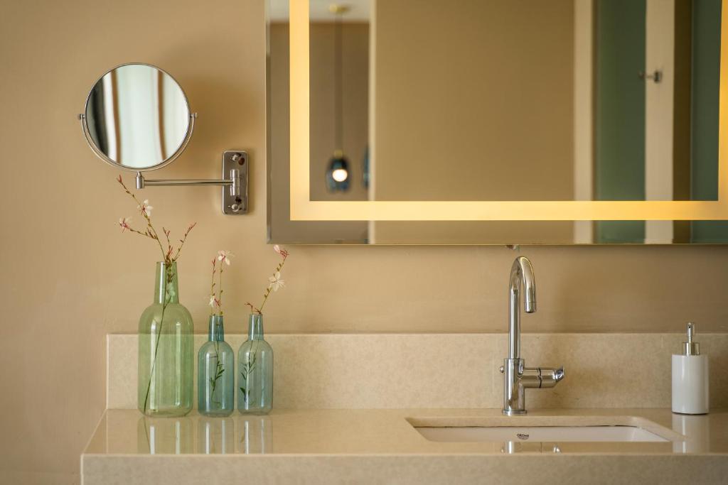 Mūsdienīgas vannas istabas interjers ar spoguli, stikla pudelēm ar ziediem un elegantu izlietni. Rāmja apgaismojums papildina telpas estētiku.