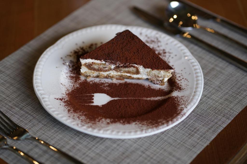 Tiramisu gabals uz baltas šķīvja ar kakao pulveri ap malām, blakus galda piederumi un galdauts.